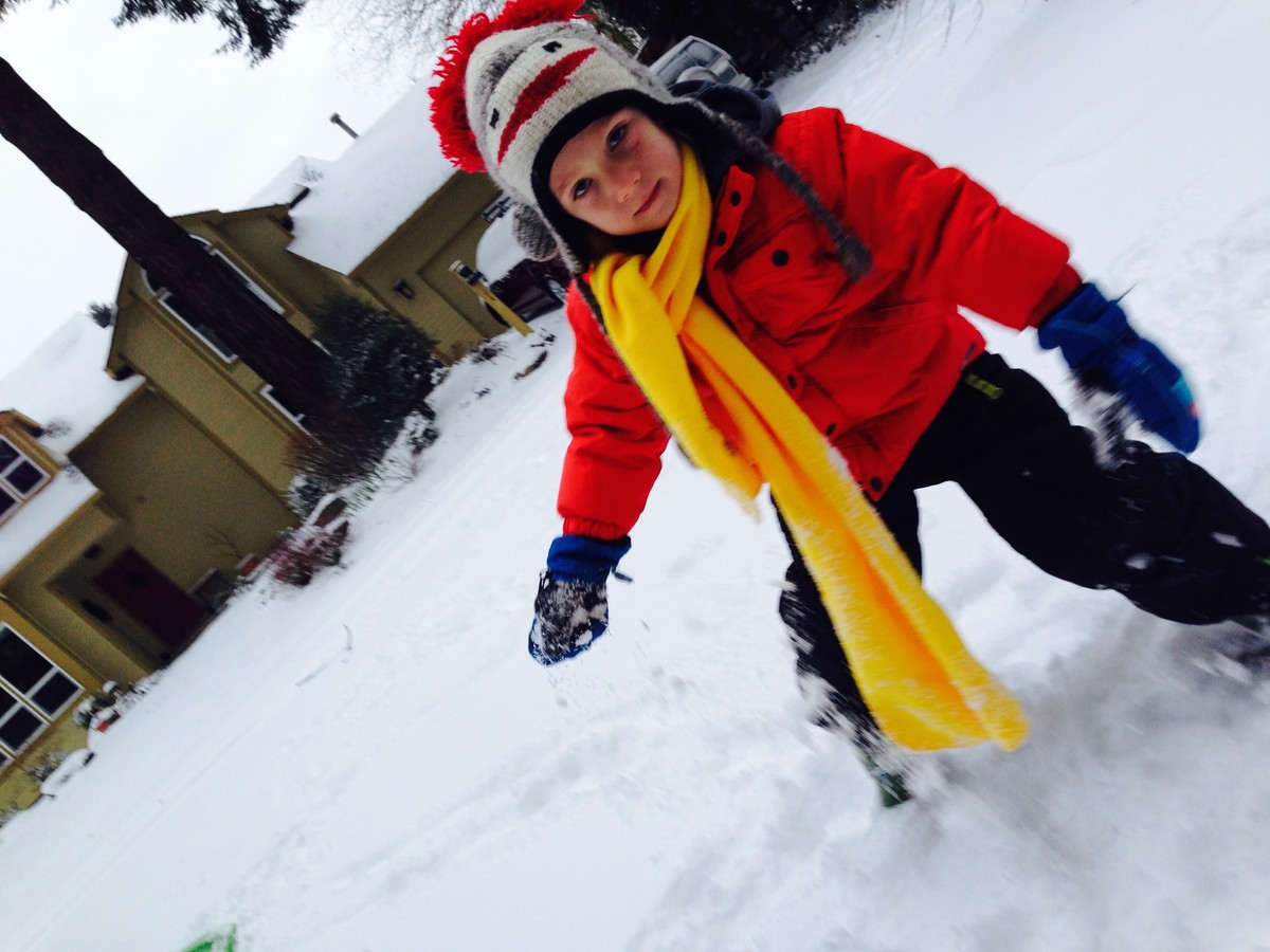 Kindergartener in colorful snow clothes in a snowy street. He has a hat styled to look like the head of a sock monkey toy