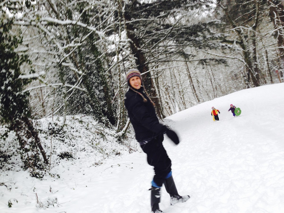 A woman at the top of on a sloping snowy street that dead-ends in a forest, looking toward the camera. Downslope two children in colorful snow clothes are running up the street (toward us) with a plastic disc sled