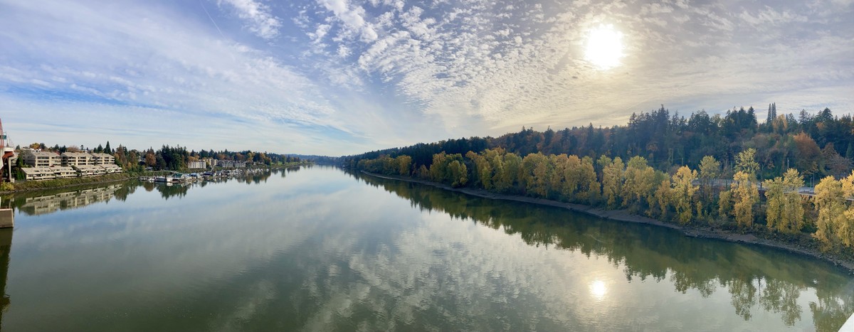 Panorama looking south up the Willamette River from a bridge about 200' above the water. The glassy river reflects a strangely greenish blue sky speckled with high altitude clouds. On the east bank is a marina, houseboats, and a condo complex. On the west bank are hills forested with dark firs and a few red maples; along the shore poplars and cottonwoods are bright yellow with fall foliage. The afternoon sun is low in the sky and shines weakly through the veil of clouds