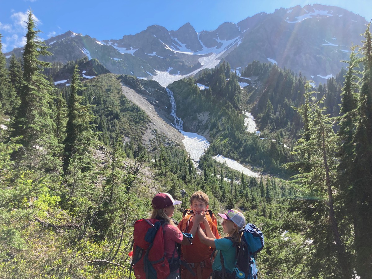 Steep mountain landscape in brilliant sunshine with three kids in backpacks. Two of the kids are using their fingers to force the third’s mouth into a smile shape. About a mile distant are three hanging valleys leading to a lightly glaciated row of peaks; a waterfall drains the topmost glaciers into an ice-covered lake in the lowest valley