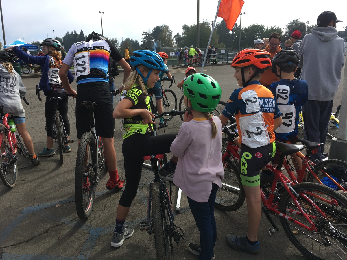 A crowd of children on bicycles, in racing gear, all facing away from the camera, staging for a race. Three kids are the focus of this shot in center frame, in conversation