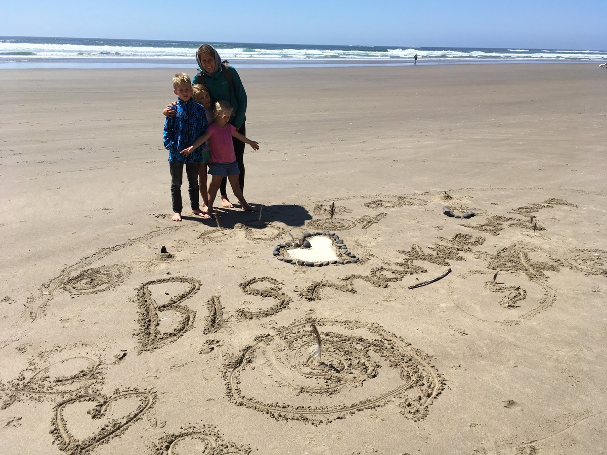 Shot on a beach of a woman and three children in front of a temporary shrine made in the sand. Reads: “Bismarck we love you” with a large heart made from Bismarck’s ashes, and other small bits from the beach like sticks, rocks, and shells