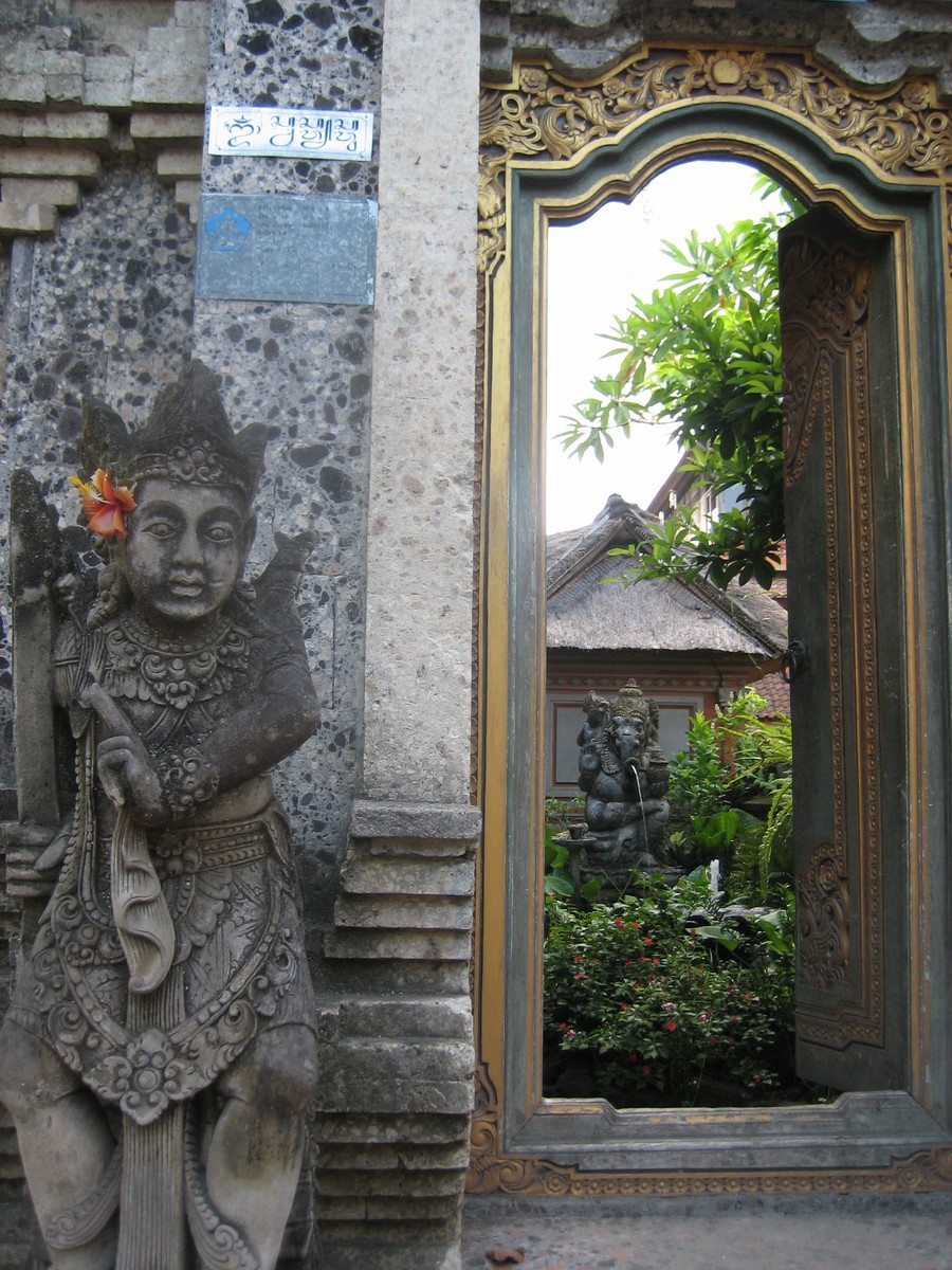A doorway in Bali: outside is a statue of a woman with a flower in her hair, inside the doorway is a courtyard with a statue of Ganesha