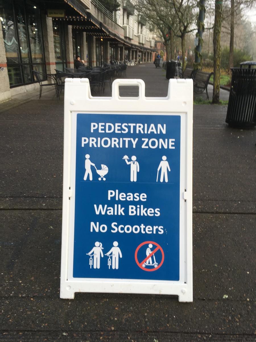 Sandwich board sign on an empty sidewalk, reading “Pedestrian Priority Zone: Please walk bikes / no scooters”