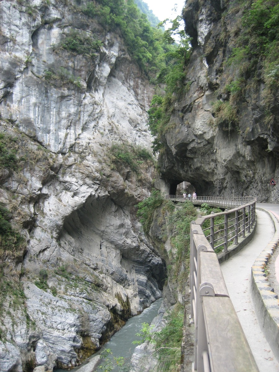 A narrow road with a railing and sidewalk, literally carved from a cliffside, in a steep narrow canyon above a small swift river. The road passes through a short tunnel about 200m ahead