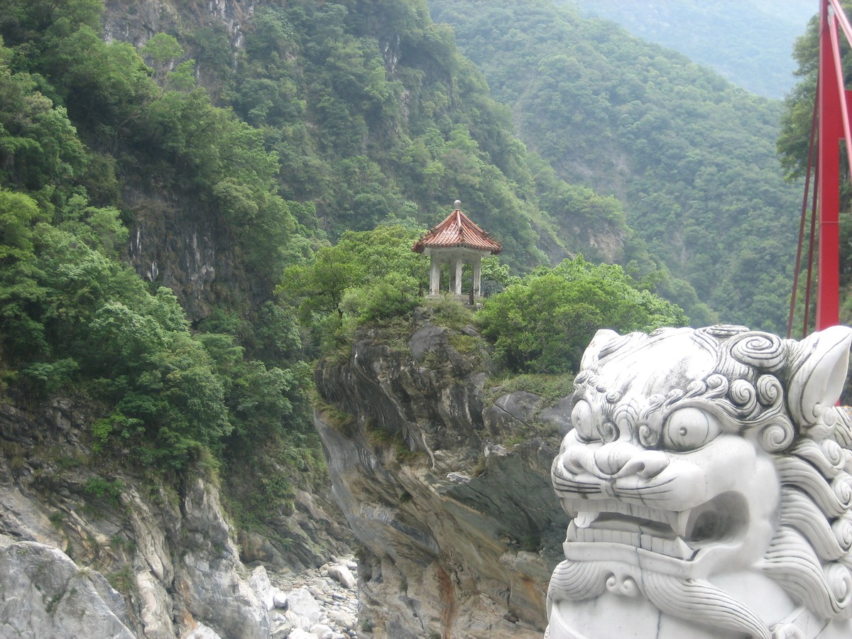 Landscape up a narrow river valley with white marble banks, steeply forested above. In the foreground, a white marble lion statue. About 200m distant, a small shrine on a rocky prominence above the river