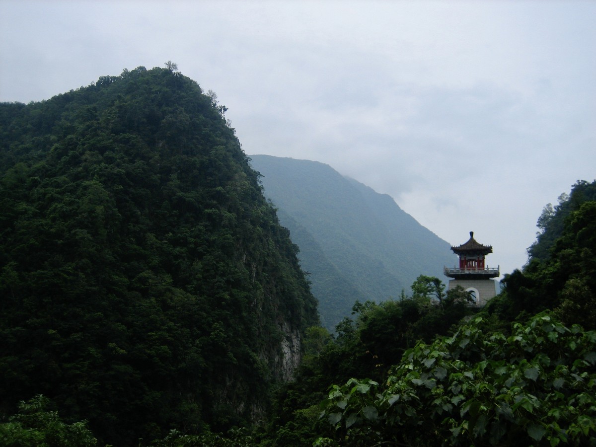 Landscape of steep forested hills, hazy with tropical moisture. A small shrine or temple is in silhouette on the side of the nearest hill, perhaps a kilometer distant