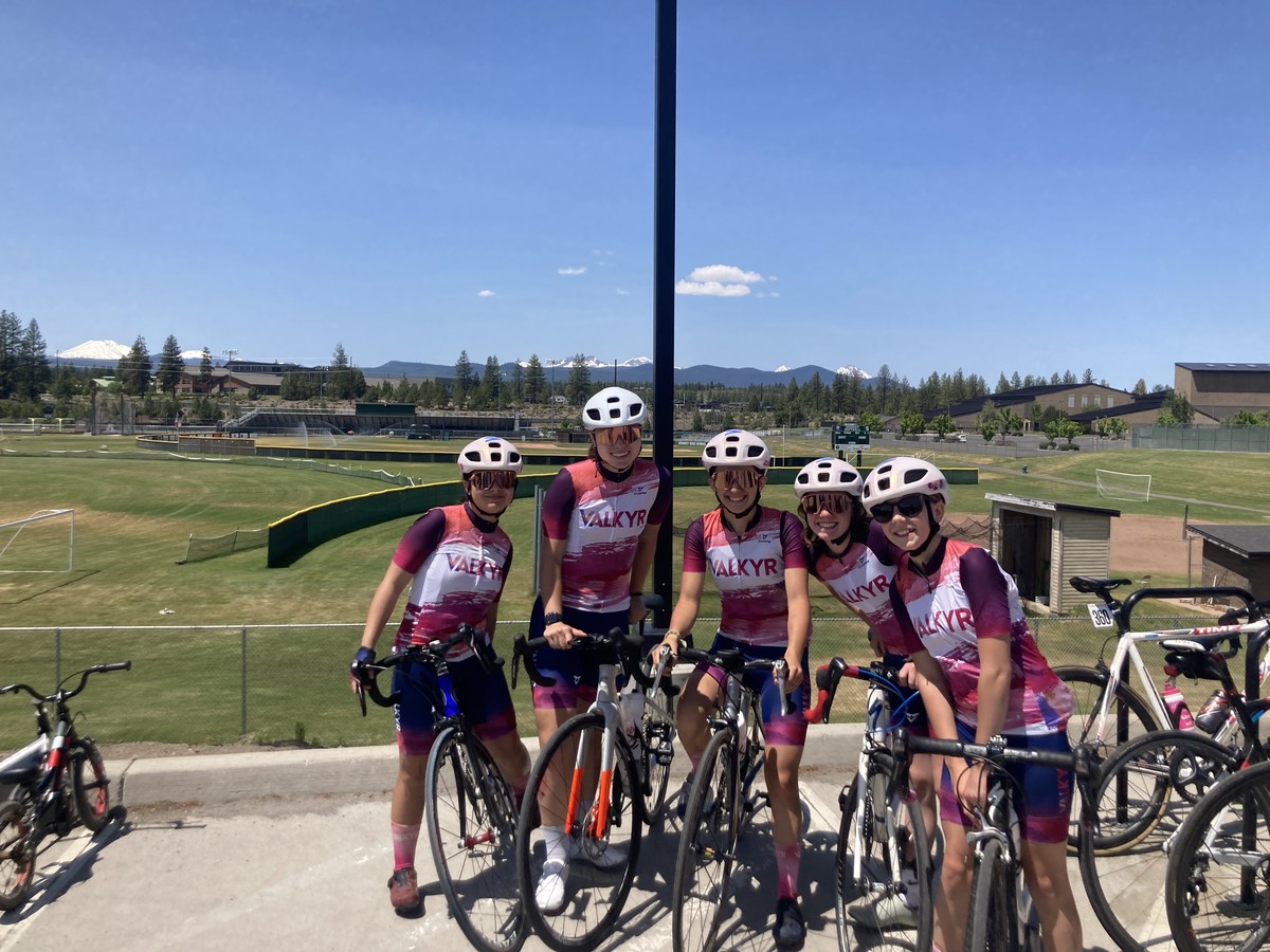VALKYR juniors in kit after Sunday ride, with bikes, at the edge of a field. Mt. Bachelor and the Sisters, snowy, are peeking over a forested ridgeline on the horizon