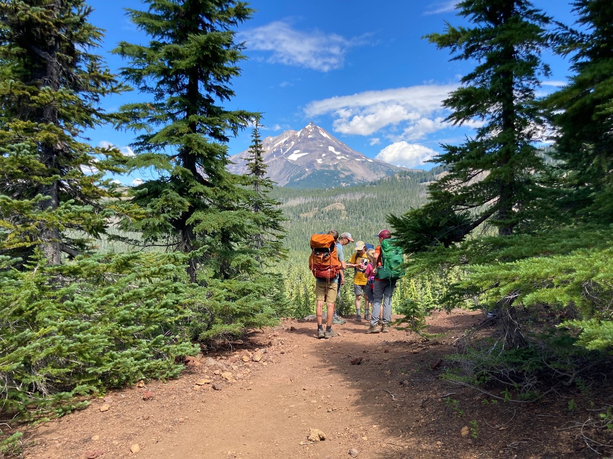 A group of hikers huddled together about 25' distant, sharing out handfuls of candy. They are on the edge of a ridge, flanked by small firs, with Mt. Jefferson on the horizon