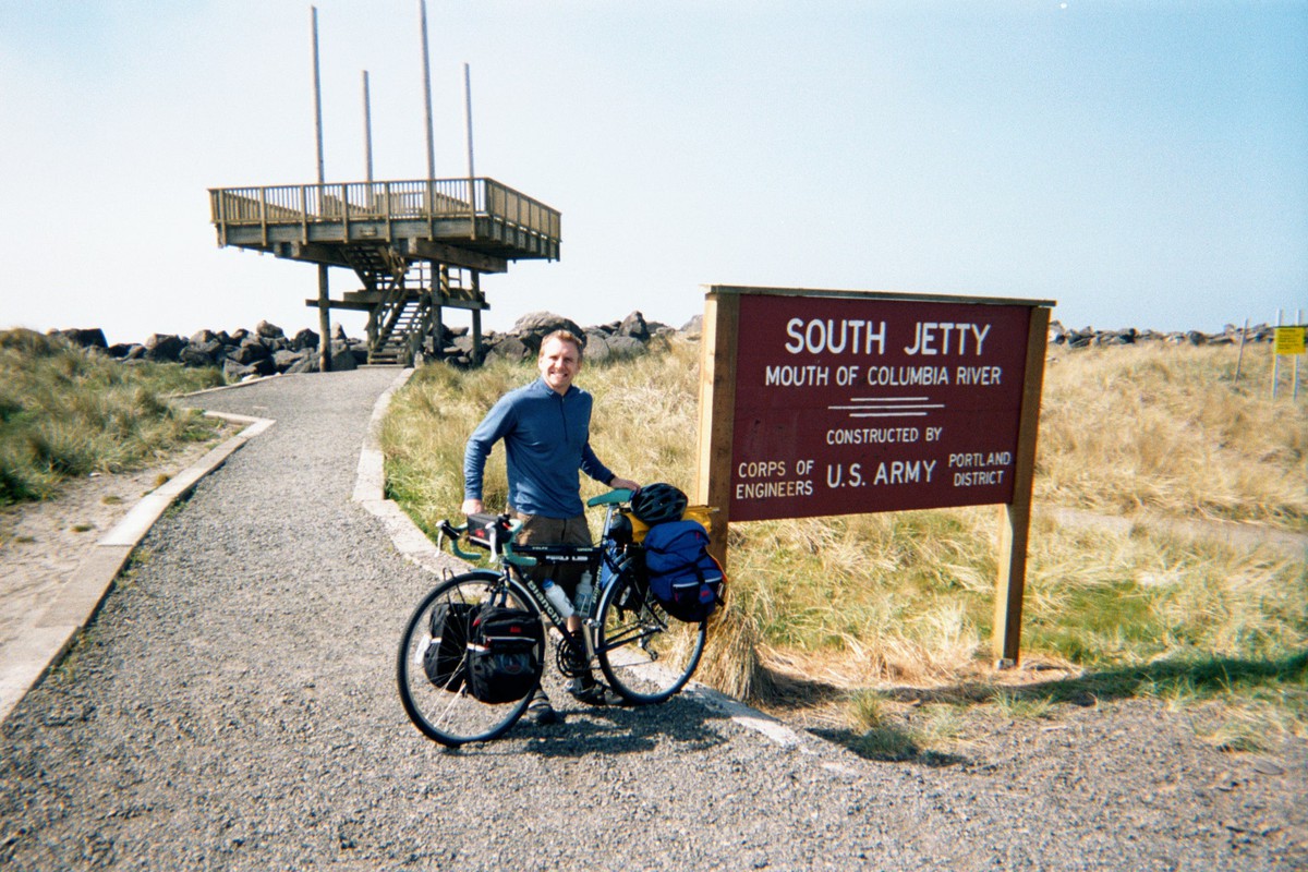 Casual portrait, outdoors, of a young man with a touring bicycle next to a sign that reads “South Jetty — Mouth of the Columbia River — constructed by U.S. Army Corps of Engineers Portland District”