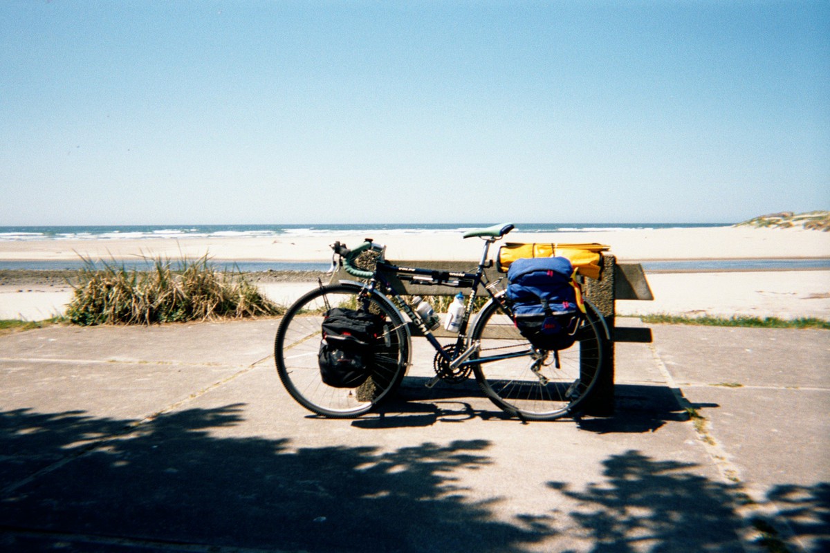 Touring bicycle, fully loaded with bags and camping equipment, leaning against a picnic table at a sunny beach