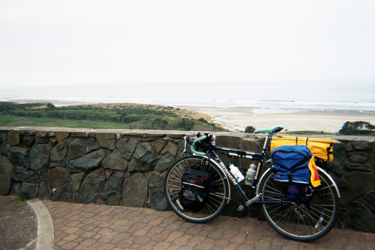 Loaded touring bicycle leaning against a low stone wall at a wayside viewpoint above a broad beach on the Pacific. Air is gray and damp.
