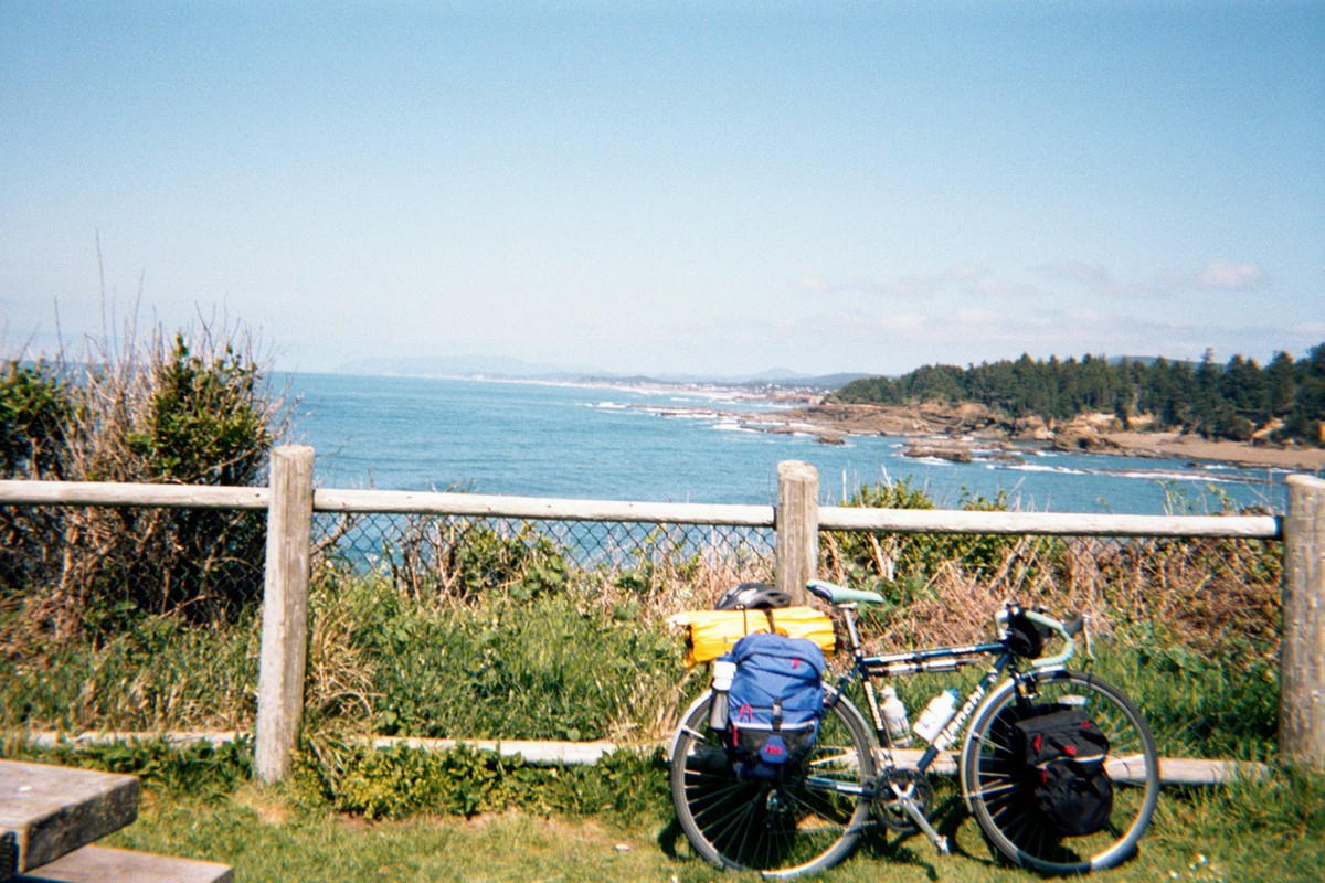 Bicycle leaning against a fence at a wayside viewpoint, looking north into a wide bay with a forested headland on the far side. Weather clear and windy