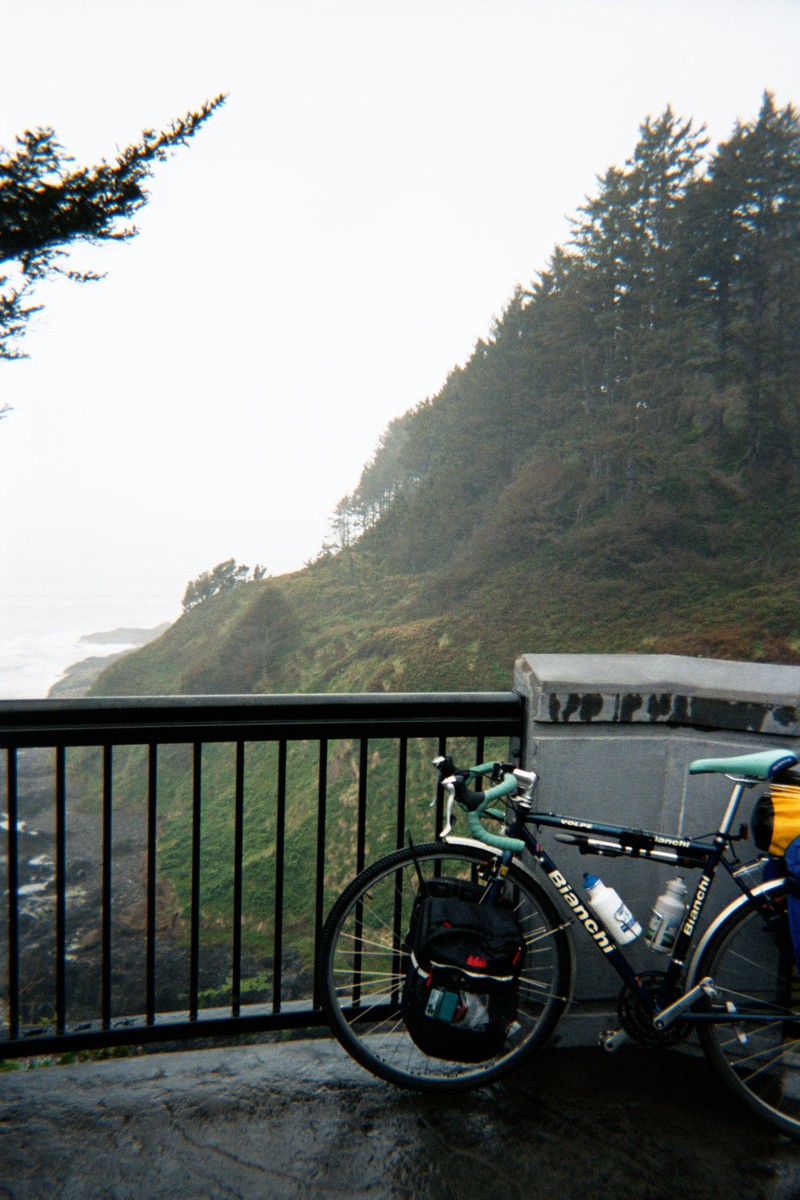 Loaded touring bicycle leaning against a metal and concrete railing on a steep, forested hillside next to a rocky beach. Weather misty and rainy.