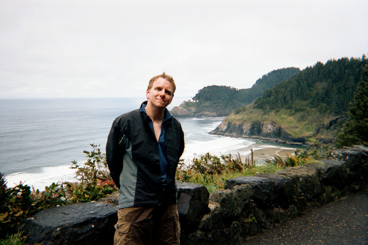 Landscape and casual portrait of a 30-year old man in wet bike gear standing at a roadside pullout on a seaside cliff with the ocean and two headlands in the background. The farthest headland is Heceta Head, with a lighthouse