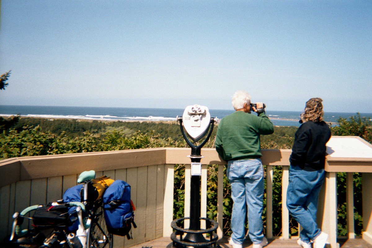 Two sightseers with binoculars on an observation platform looking west, away from the camera, across a broad grassy dune toward the ocean. In the foreground a heavily loaded touring bike rests against the railing