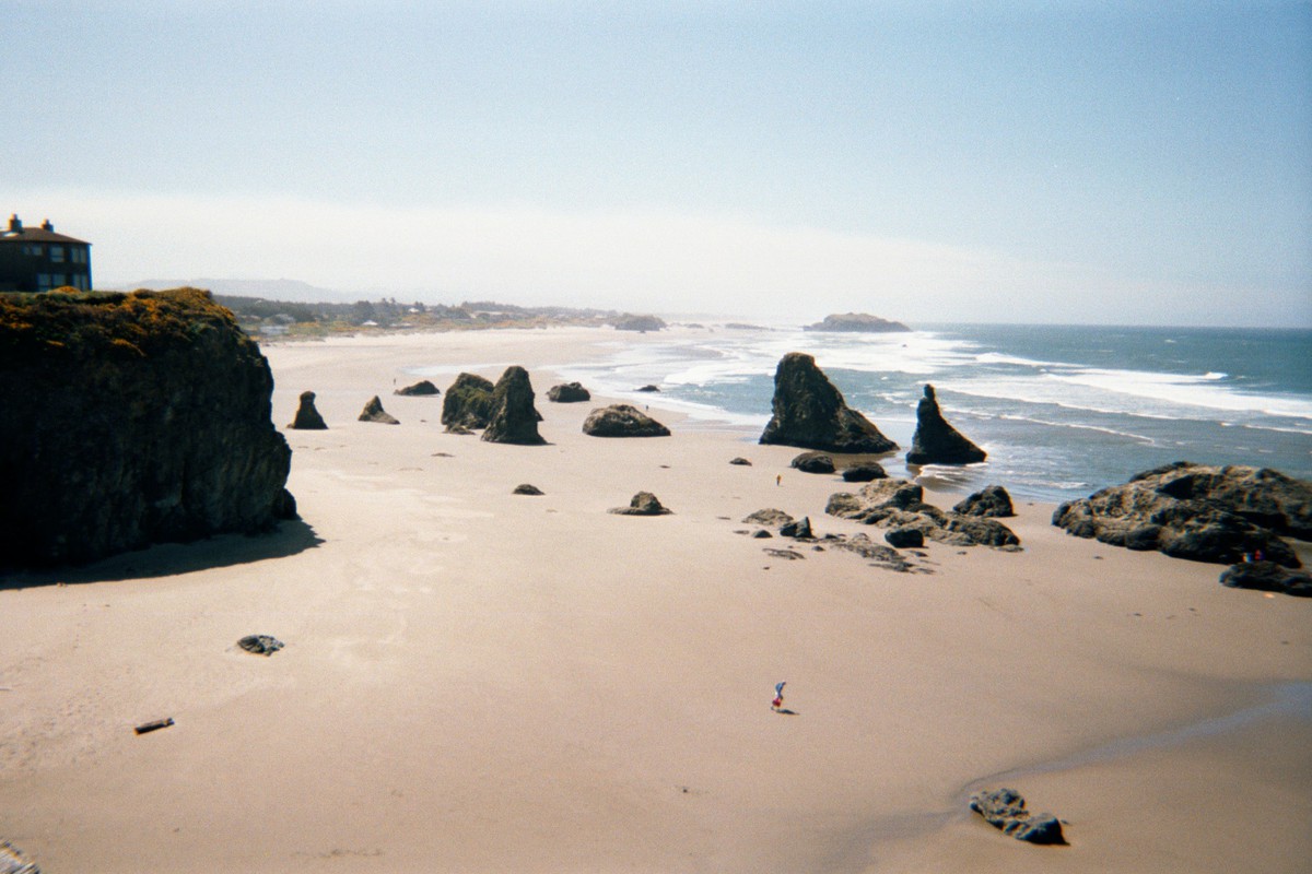 A broad beach with a dozen or so tall large (20' tall) rocks. A single person is in the center of frame, tiny in comparison. Weather sunny, windy