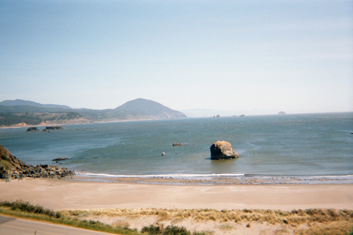 Landscape over a narrow beach & across a broad bay with scattered seamount. On the horizon, a prominent hill or low mountain projects into the ocean. Weather sunny and windy