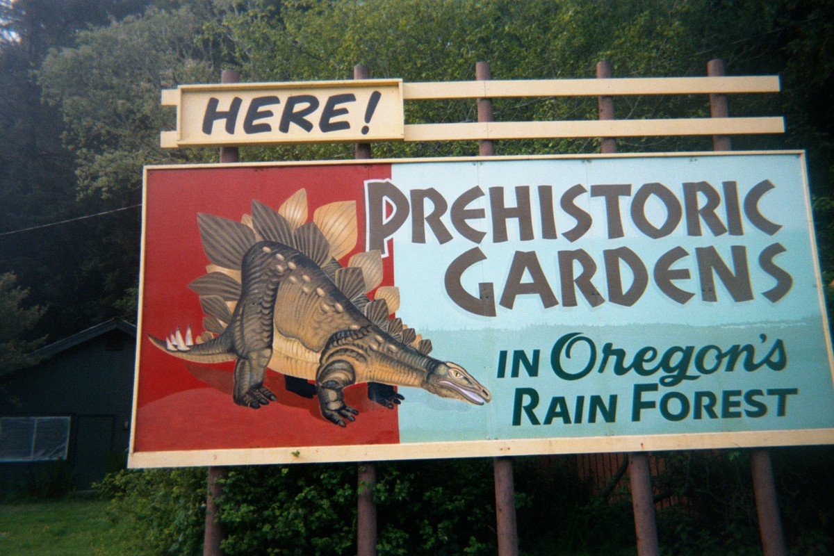 Colorful billboard with an illustration of a Stegasaurus. Text reads “HERE! PREHISTORIC GARDENS in Oregon’s RAIN FOREST”