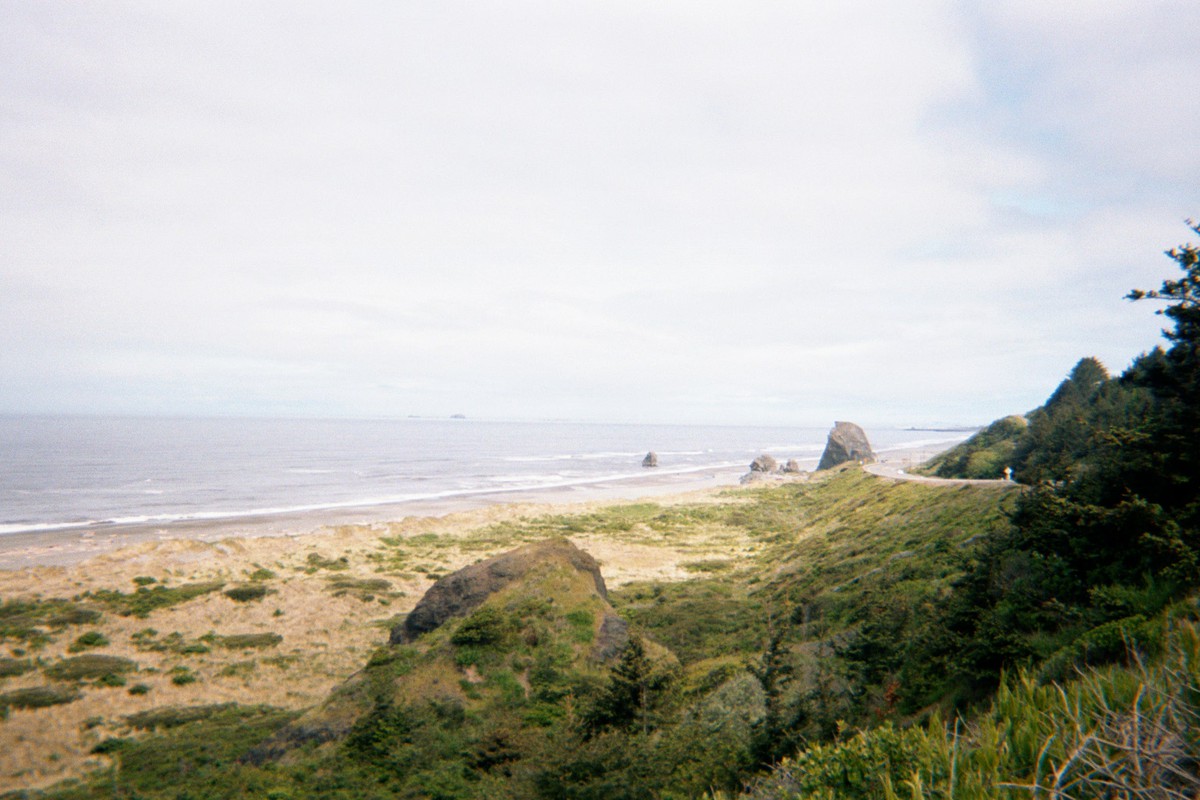 Sea/landscape facing north with ocean to the right. Narrow sandy beach grading into a steep hillside with a nearly-empty highway wending along it. Weather mostly cloudy, windy