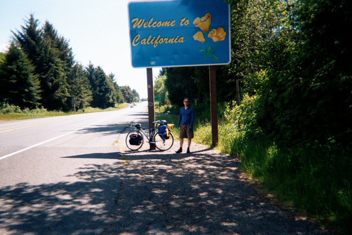 Nearly empty highway in the woods. 30-year-old white man in baggy bicycle clothes standing next to a loaded touring bike leaning against a sign that reads “Welcome to California.” Weather sunny