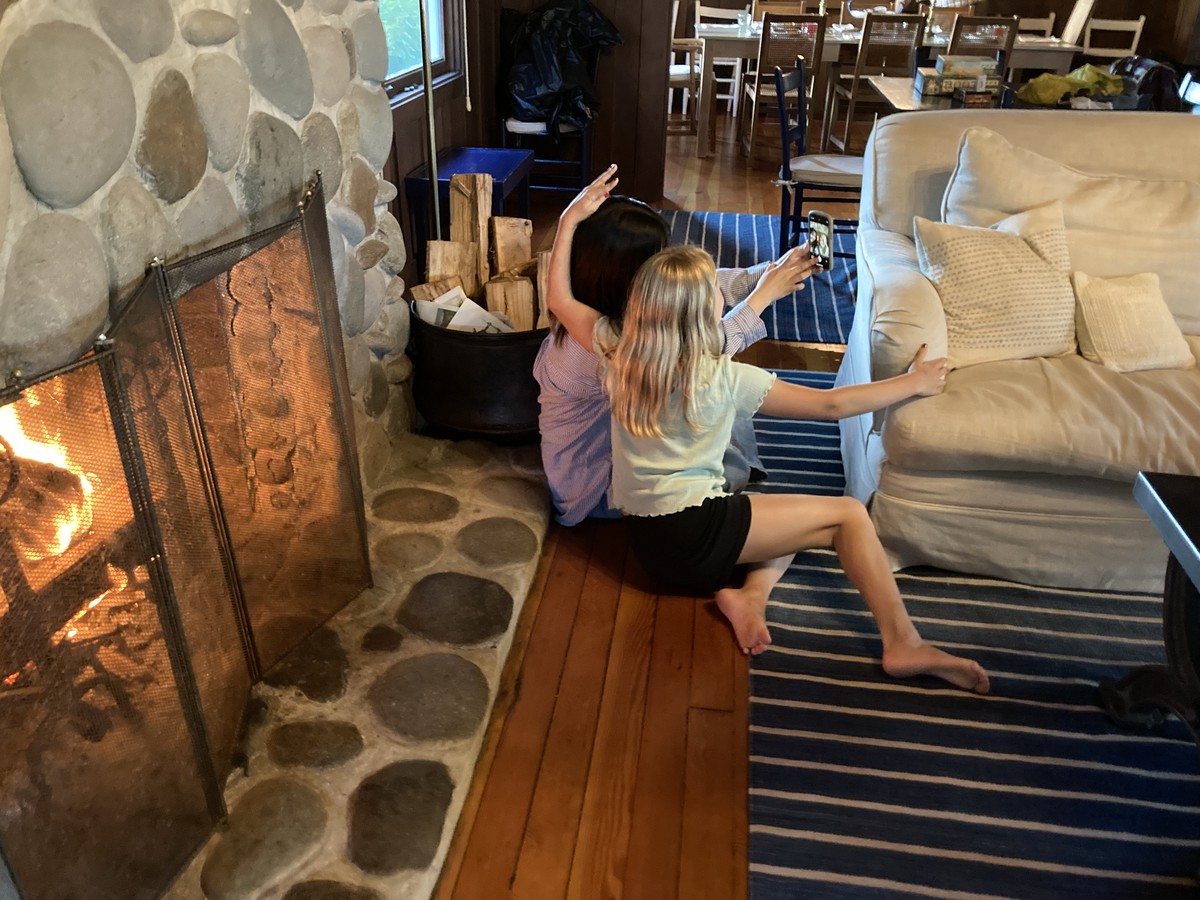 A girl an a young woman seated in front of a fireplace, taking a selfie, shot 3/4 from behind