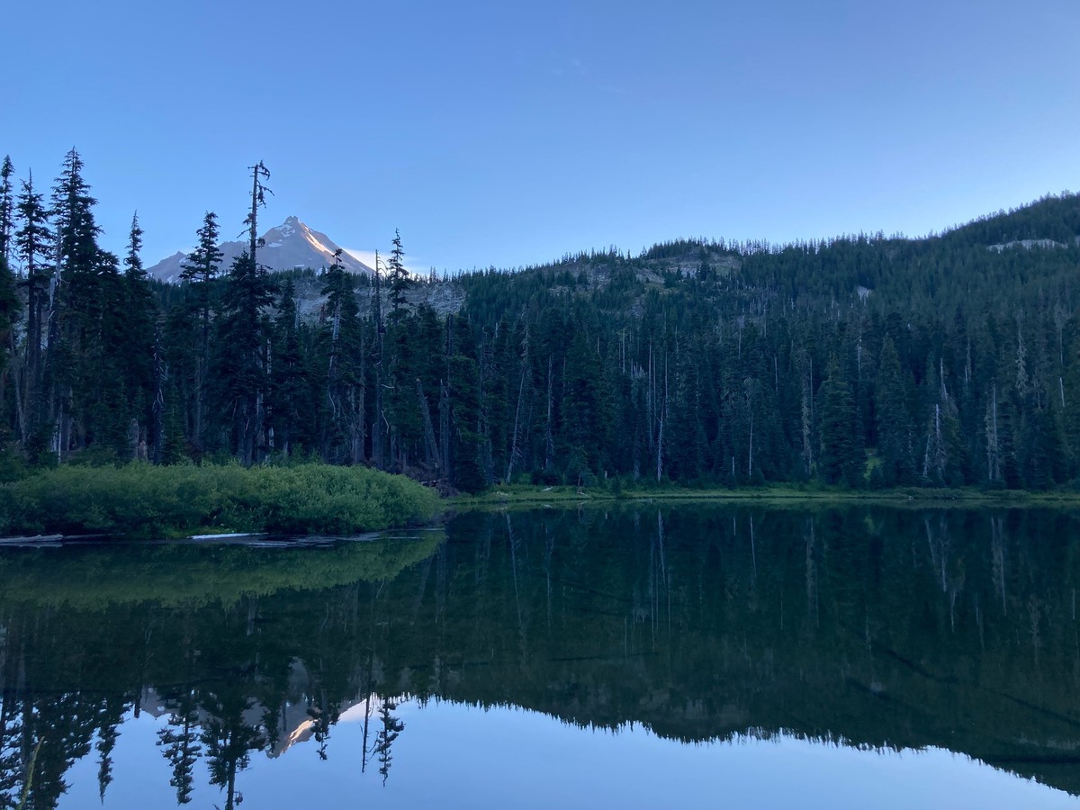 A mirror-still alpine lake at sunrise, in cool shadow, with sunlight just striking the peak of Mt. Jefferson above it