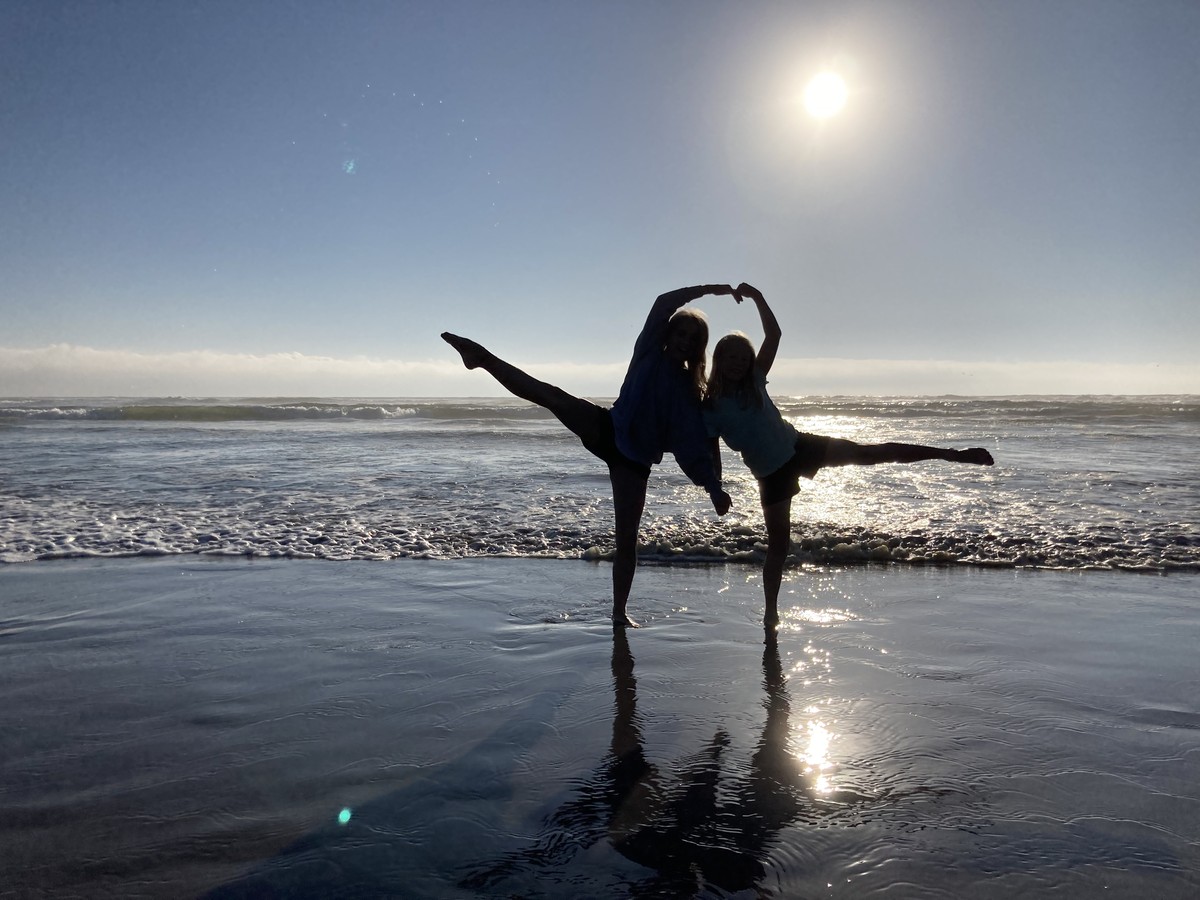Two school-age girls in silhouette making a symmetric Arabesque in shallow water, with the sun setting behind them