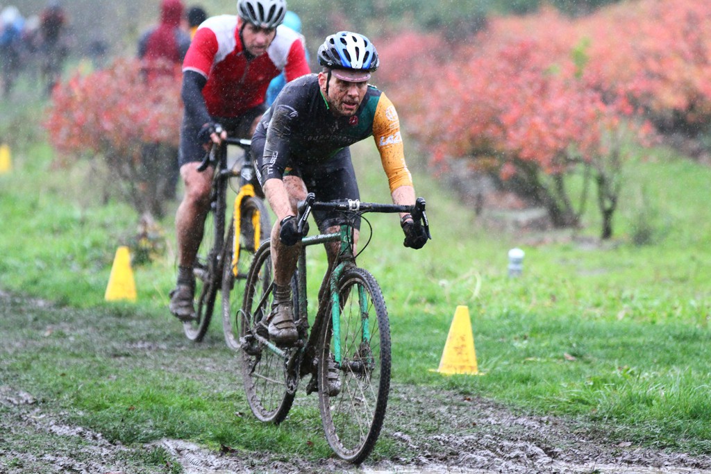 Wet and muddy cyclocross bicycle racers cycling past blueberry bushes in a heavy rain