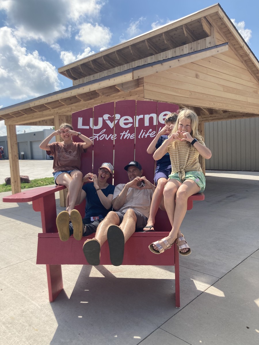 Two adults and three school-age kids sitting in a comically-oversized rocking chair placed especially for tourist photos, on which is written “Lu❤️erne: love the life!” All five people are making heart symbols 🫶 with their hands