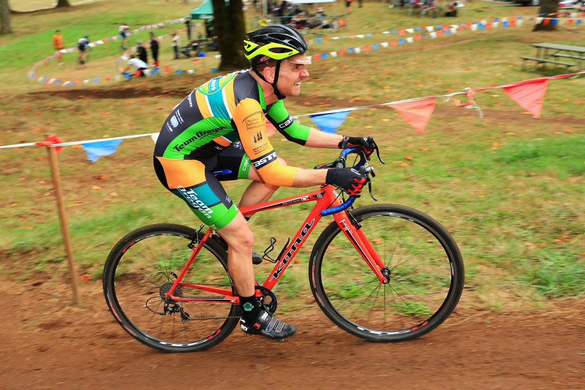 Middle aged man in Team Oregon skinsuit on a cyclocross racing bike, face contorted with visible effort