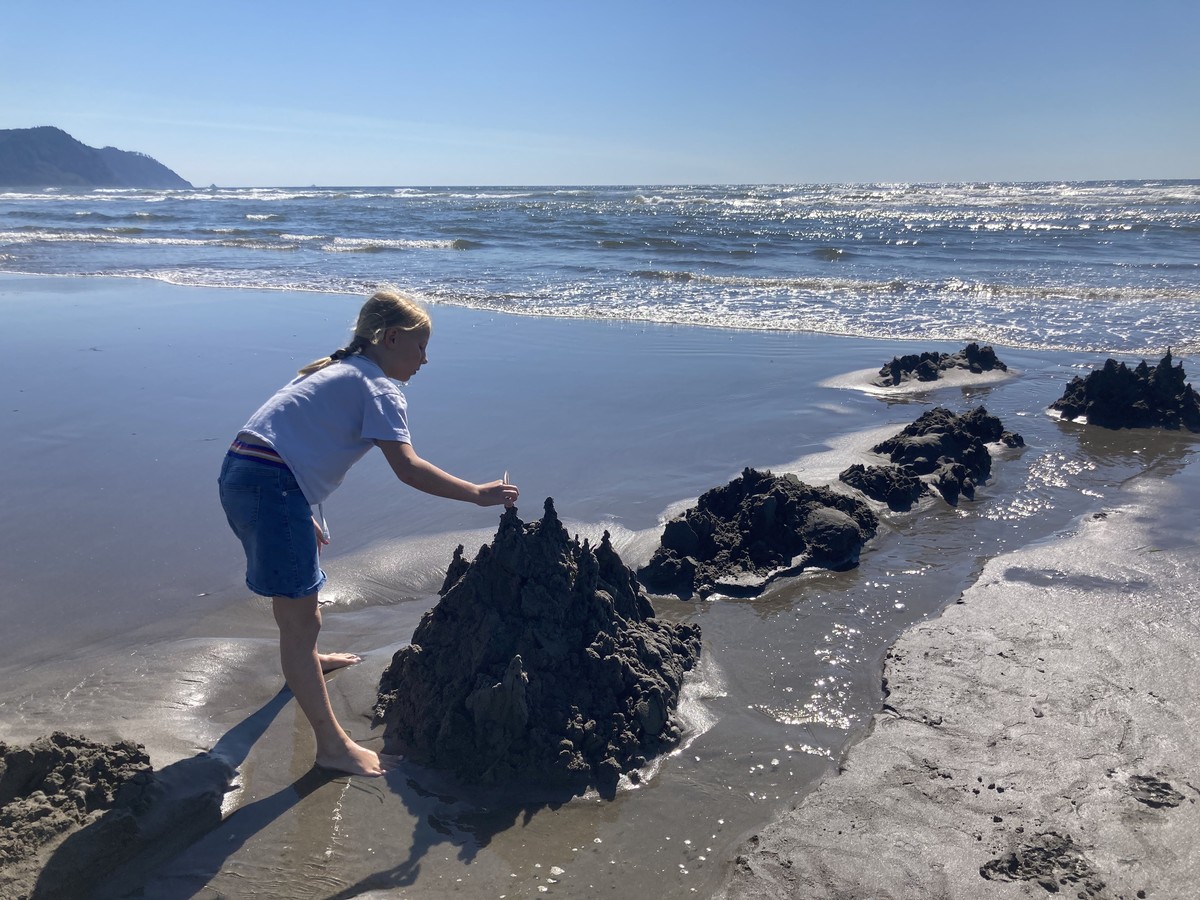 School-age child on an Oregon beach making a “drip sandcastle” with her bare hands, under a very clear July sky