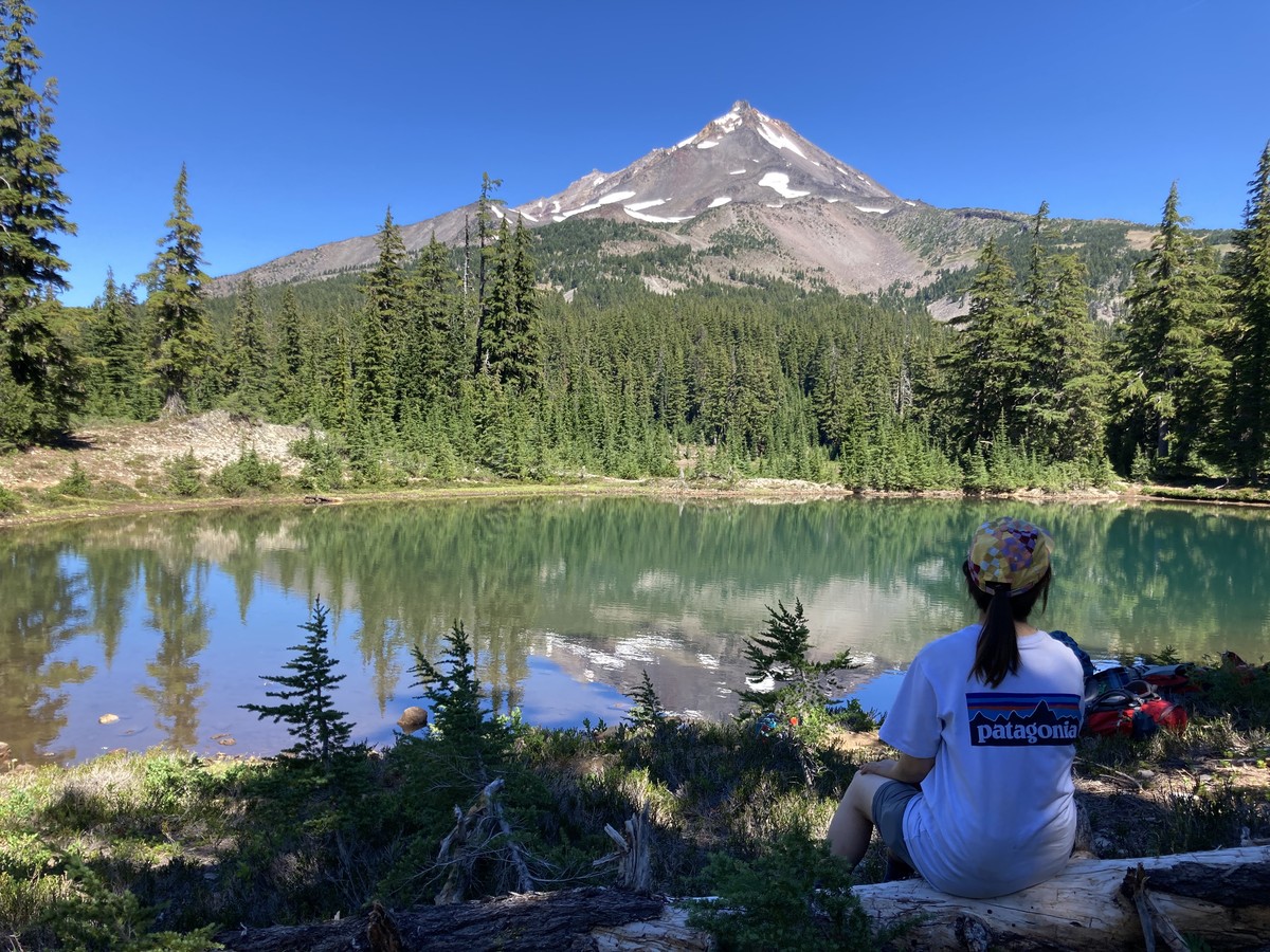 A twenty-something woman seated on a log in the shade next to a small alpine lake, looking toward the peak of Mt. Jefferson which is nearly snow-free