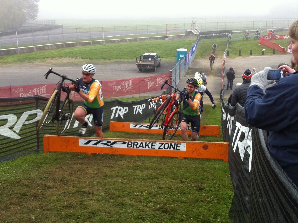 Shot downhill at a muddy cyclocross race. Two middle aged men are hoisting their bikes over a series of barriers