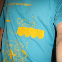 Selfie of a torso with a bright aqua t-shirt with a simplistic cartoon of a band underwater next to a dog above water. Text reads The Young Fresh Fellows