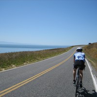 Outdoors, taken from a moving bicycle: a narrow empty highway along the seashore; a young woman on a road bicycle just ahead and moving our direction. Brilliant clear sunny weather