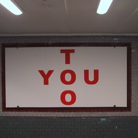 Simple billboard in a Berlin u-bahn station, reading “YOU TOO” in large block letters such that the Os in both words intersect, forming a cross shape