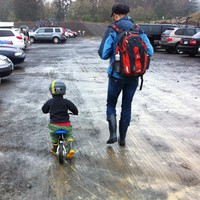 Woman and preschooler in a parking lot at a gravel quarry. Both are wearing rain gear and rubber boots. The child is riding a pedal-less scoot bike