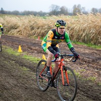 Middle-aged man on a cyclocross racing bicycle, wearing a colorful racing skinsuit, riding down a muddy track next to a field