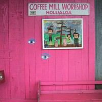 Empty bench in front of a colorfully-painted wooden barn, sign reads “Coffee mill workshop, Holualoa”