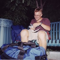 23-year-old white man, slightly drunk seated on a bench with a large duffel bag at his feet. Taken late at night, with a flash. He is looking at a train ticket