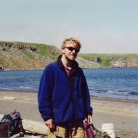 Casual portrait of a disheveled young man with a beard and sunglasses, wearing a dingy fleece jacket, holding a mason’s trowel. Taken on a sunny day on beach in the subarctic, with green cliffs across a narrow bay in the background