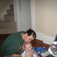 Indoors, a woman seated on the floor, kissing a baby on his head. Baby is wrapped in a towel. In the foreground a dog’s snout is visible just in the edge of the frame