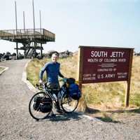 Casual portrait, outdoors, of a young man with a touring bicycle next to a sign that reads “South Jetty — Mouth of the Columbia River — constructed by U.S. Army Corps of Engineers Portland District”