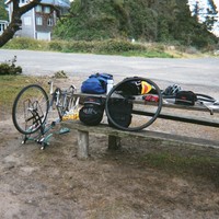 Picnic table next to a small parking lot at a coastal wayside area. A bicycle is upsidedown on its handlebars next to the table. All the touring gear is spread across the table; one wheel is off the bike, and the tire is off the wheel.