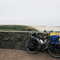 Loaded touring bicycle leaning against a low stone wall at a wayside viewpoint above a broad beach on the Pacific. Air is gray and damp.