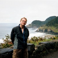 Landscape and casual portrait of a 30-year old man in wet bike gear standing at a roadside pullout on a seaside cliff with the ocean and two headlands in the background. The farthest headland is Heceta Head, with a lighthouse