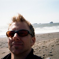 Selfie, white man with bikey sunglasses, on a windy beach facing west. Sea stacks a few miles offshore.