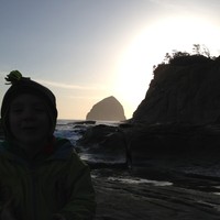 Young child facing the camera on a stormy (but clear) Oregon beach in winter. The sunset is blocked by a headland and sea stack; objects in the foreground are barely discernible as dark lumpy objects