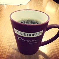 Closeup of a coffee cup on a conference room table. The cup is branded “Mercy Corps: Be the Change.” It has a Label Baby Label that reads “PAUL'S COFFEE”